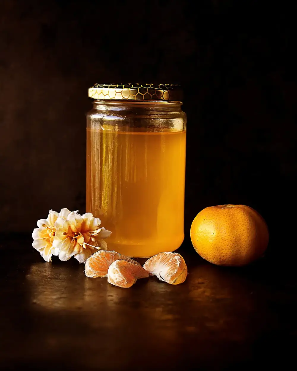 Un frasco de vidrio con miel clara y un trozo de panal encima, junto a una mandarina pelada y una flor blanca, sobre una mesa oscura