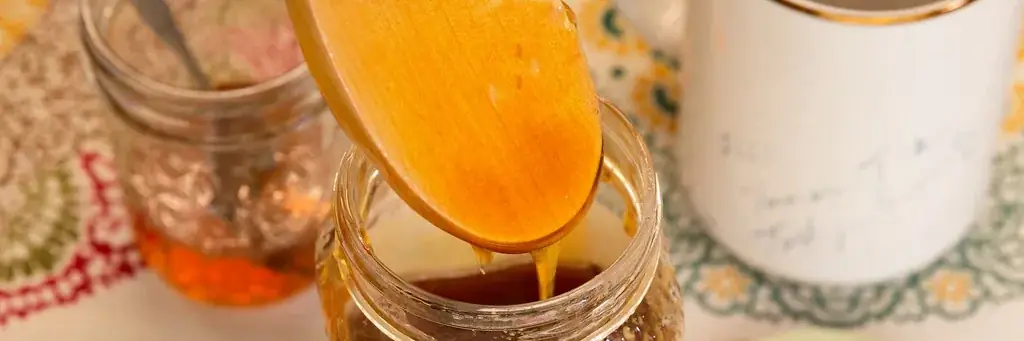 Una cuchara de madera vierte miel dentro de un frasco de vidrio, sobre un mantel bordado